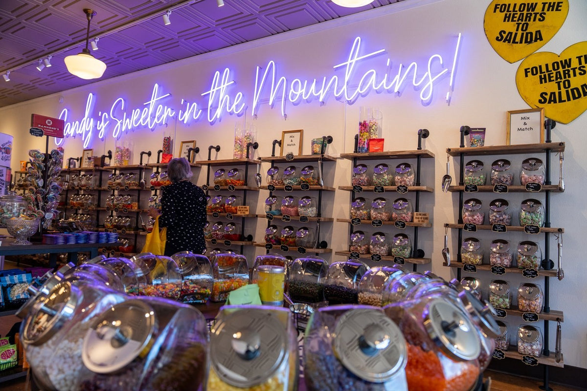 Open jars of bulk candy in front of a wall of jars under a neon sign that reads Candy is Sweeter in the Mountains