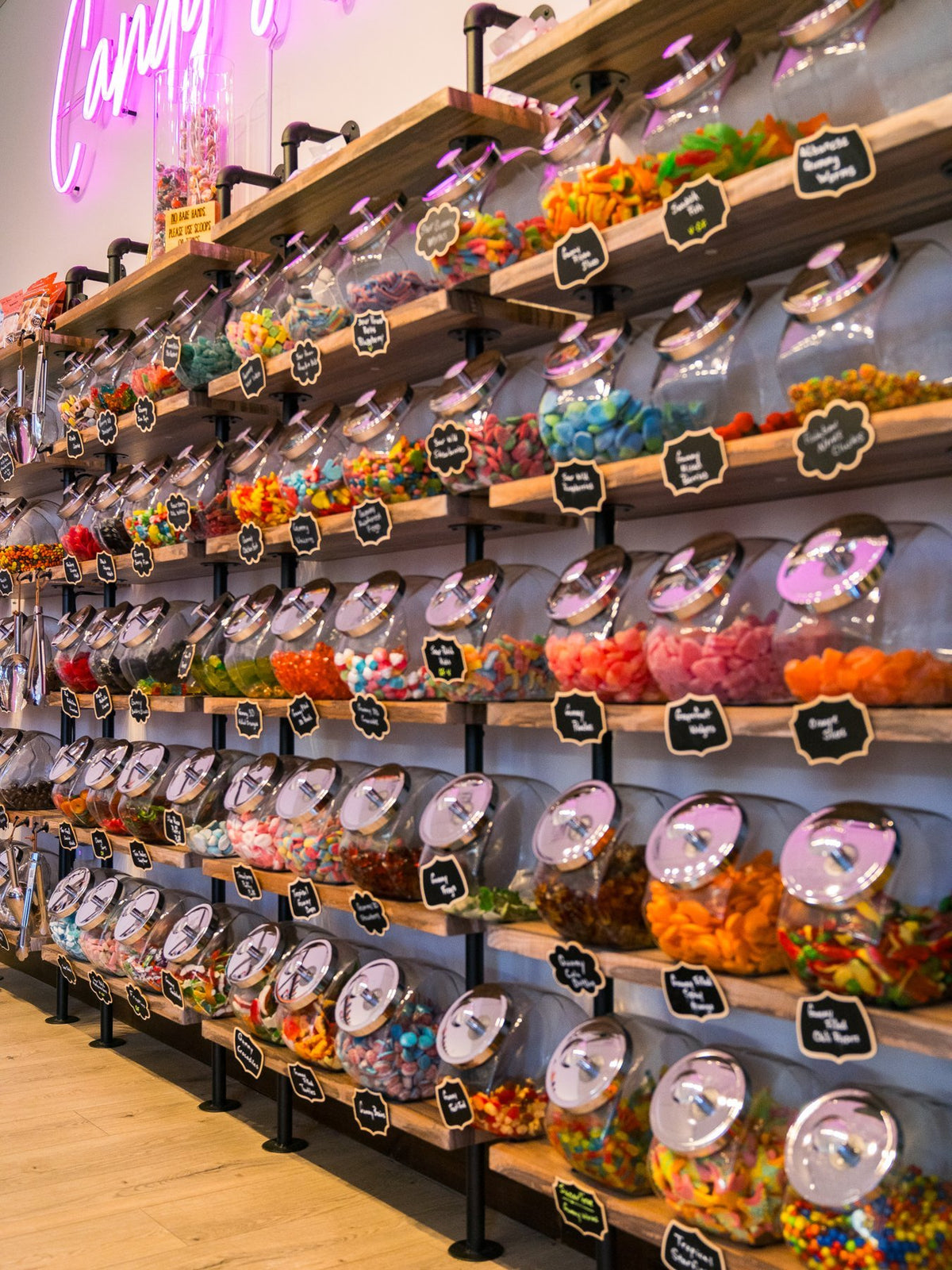 A long wall of bulk candy jars with chalkboard labels on pipe shelving